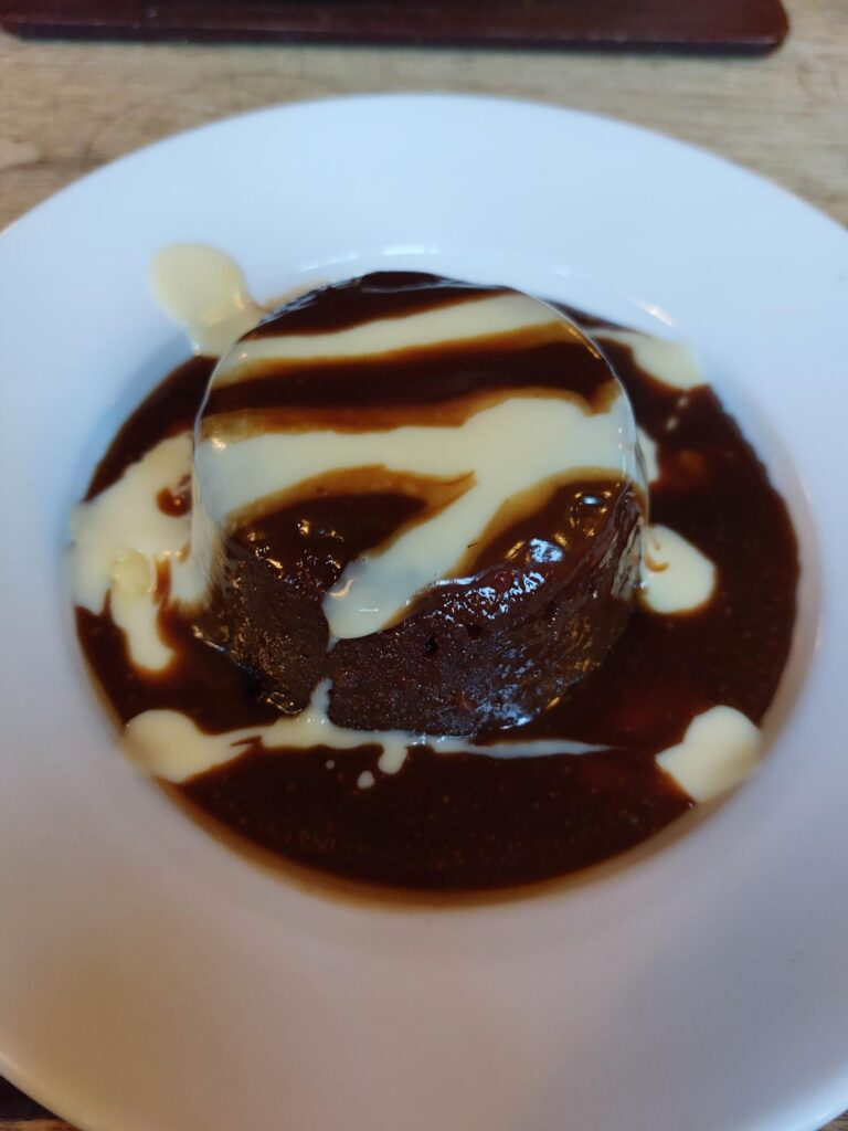 A round sticky toffee pudding covered in dark toffee sauce, drizzled with custard