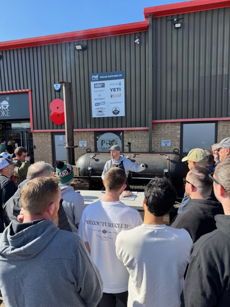 Picture of bbq class being hosted outside the shop with 500 gallon offset smoker in background with a crowd of people in front